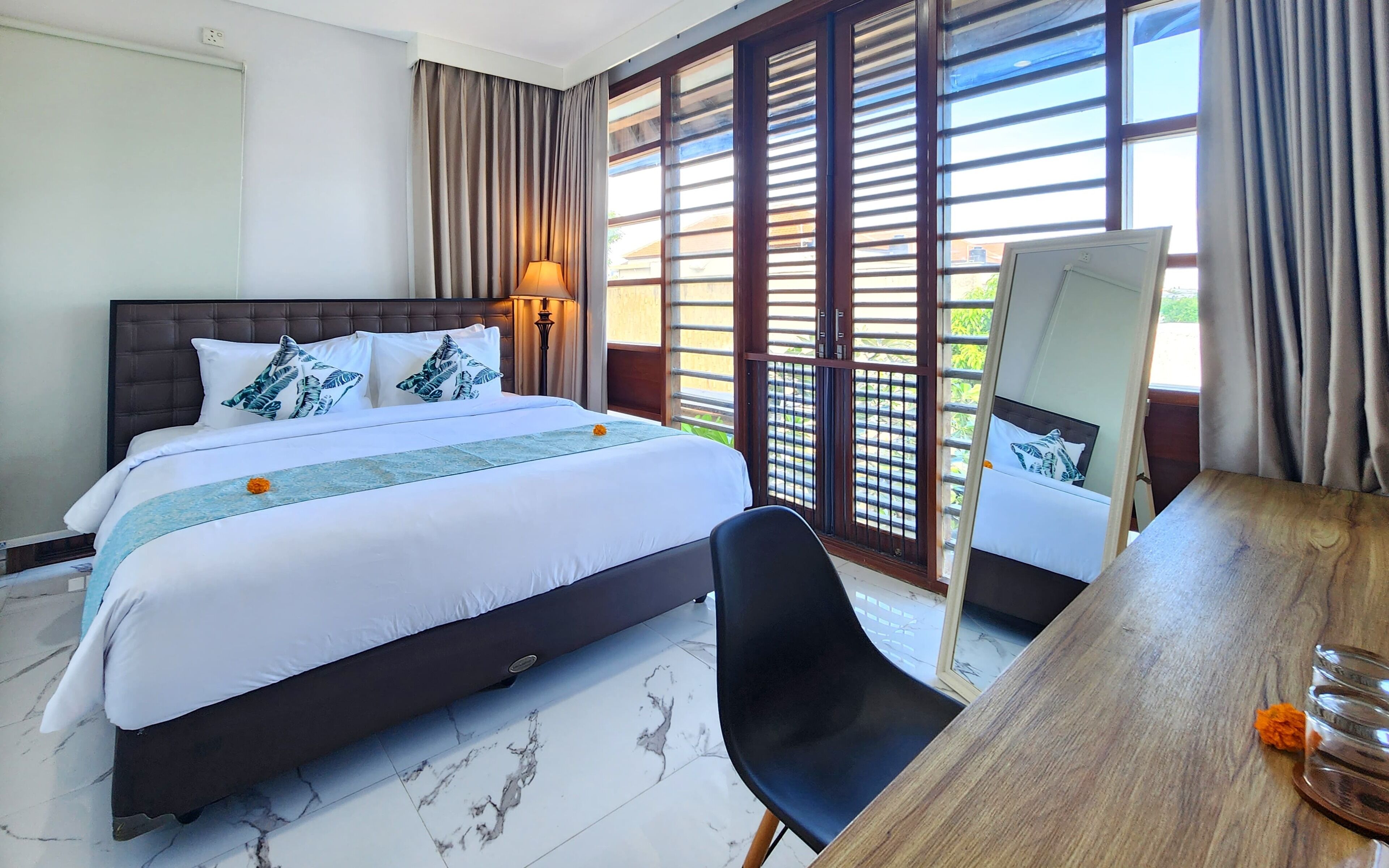 Habitación de hotel con cama, escritorio, silla, espejo y ventanas grandes con cortinas.