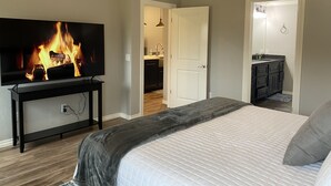 A bedroom with a bed, a TV displaying a fireplace, a bathroom, and a kitchen area.
