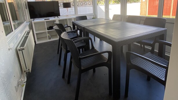 Uma varanda coberta com mesa e cadeiras, uma televisão e vista para o bairro.