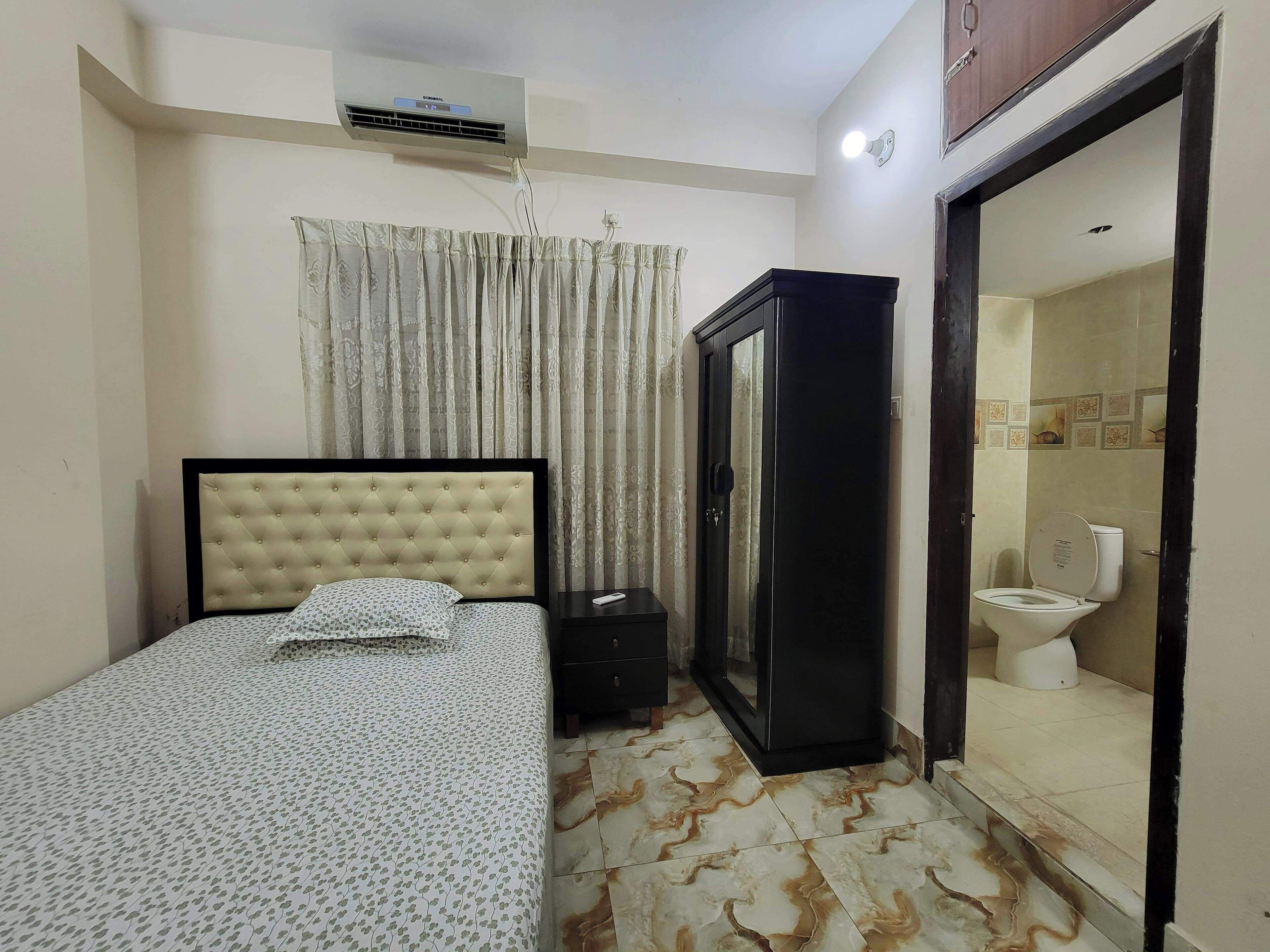 A bedroom with a bed, a wardrobe, an air conditioner, and a bathroom visible through an open door.