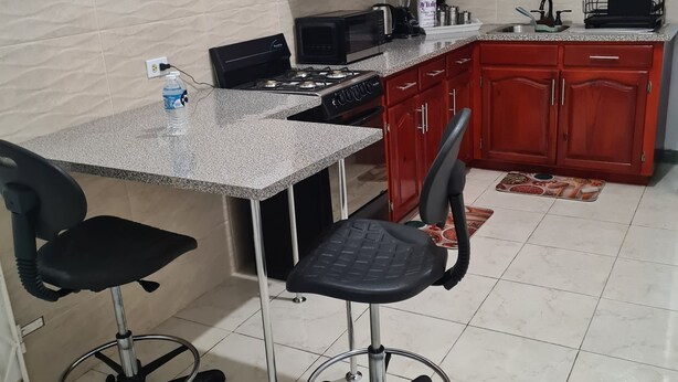 A kitchen with a granite countertop, two black office chairs, a gas stove, a microwave, and wooden cabinets. - Beautiful 2-Bed Apartment in Montego Bay (Montego Bay)