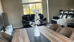 Ein Essbereich mit einem langen Tisch, schwarzen Holzstühlen, einer weißen Vase mit Blumen und einer schwarzen Couch im Hintergrund.