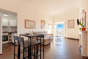 A modern living room with a sofa, dining table, and chairs, a kitchen area with a stove, and a balcony with a view.