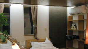 Une chambre d’hôtel avec deux lits, une table de chevet avec une lampe, une plante, une fenêtre avec des rideaux et une étagère avec des objets décoratifs.
