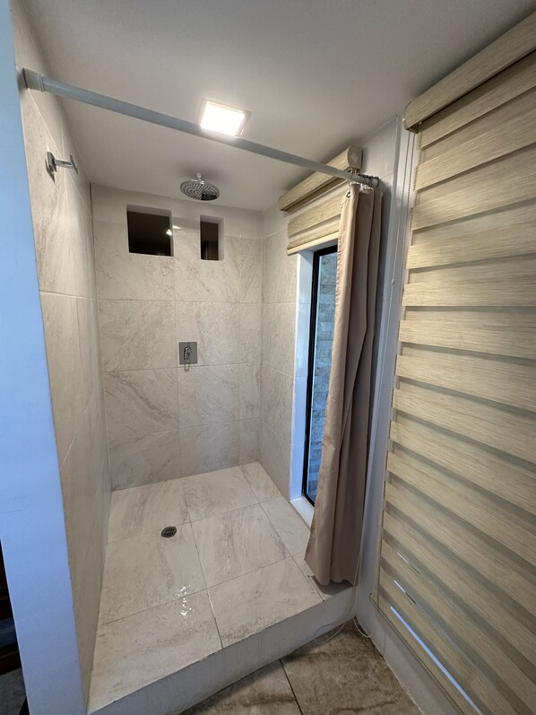 A walk-in shower with tiled walls, a ceiling light, a window with a curtain, and a metal rod for hanging items.