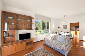 A living room with a TV, wooden shelving, a patterned sofa, and a dining area with a table and chairs.