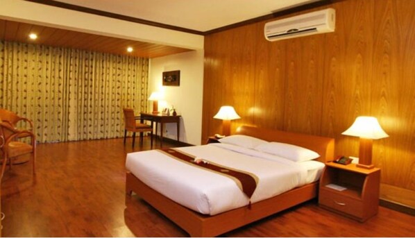 A hotel room with a wooden bed, two bedside lamps, a desk with a chair, and an air conditioning unit on the wall. - Nazimgarh Garden Resort (Sylhet)