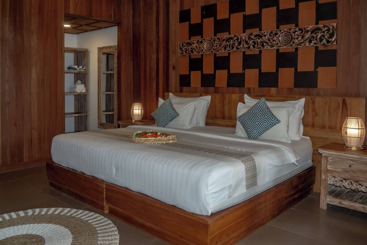 A large bed with white linens, a woven tray, and patterned pillows. There is a wooden headboard with a geometric design, a nightstand with a lamp, and a wooden floor.