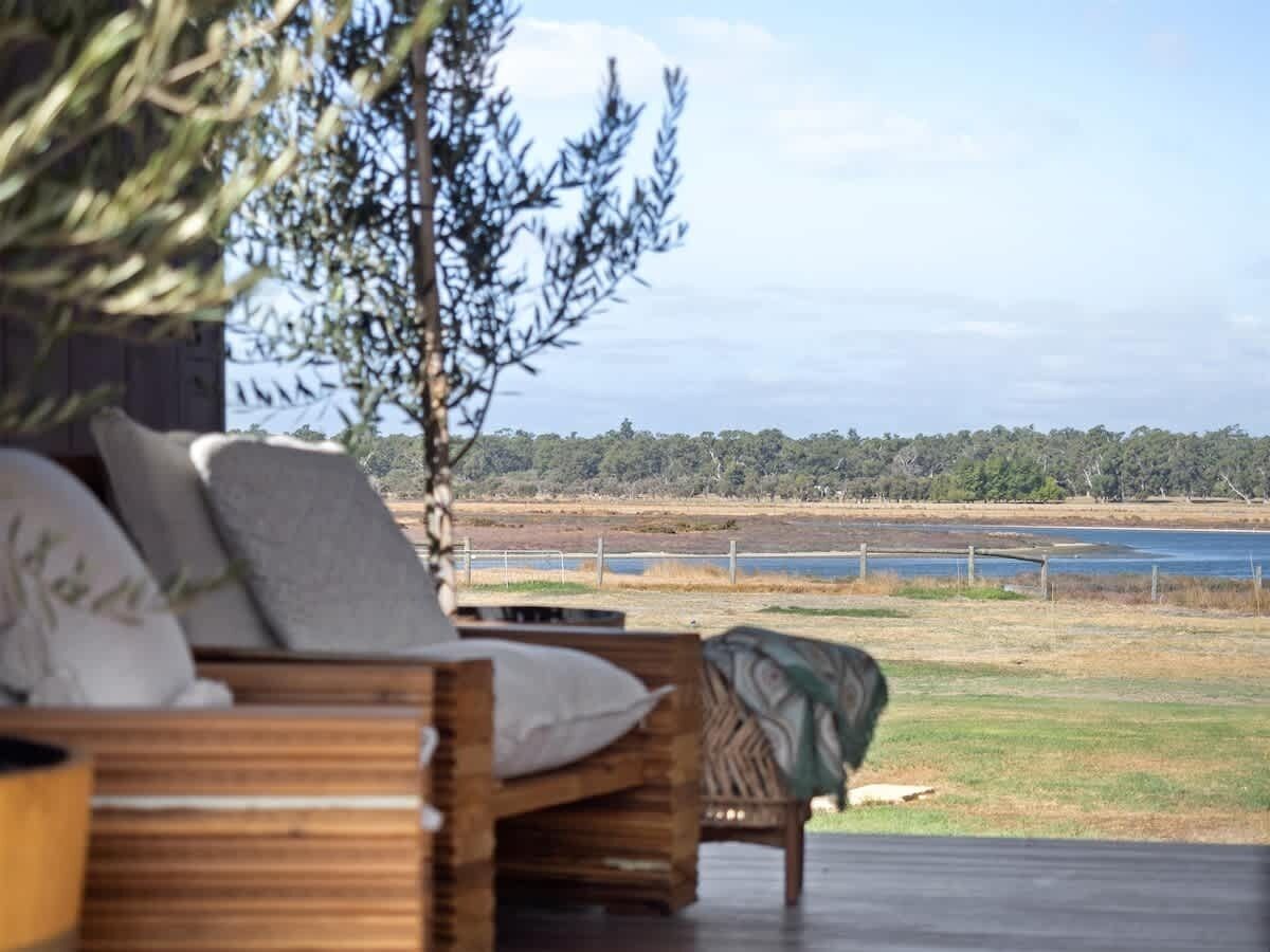 A wooden outdoor seating area with cushions, overlooking a body of water and a grassy landscape.