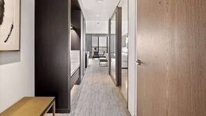 Un couloir d’hôtel avec une porte en bois, un tableau encadré au mur et une vue sur une chambre avec un lit et une fenêtre.