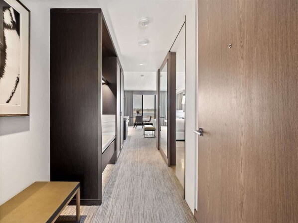 A hotel corridor with a wooden door, a framed picture on the wall, and a view into a room with a bed and a window.