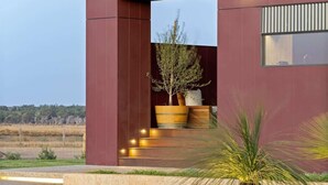 Un bâtiment moderne à la façade rouge, avec un escalier menant à une porte en bois et un pot de fleurs au premier plan.