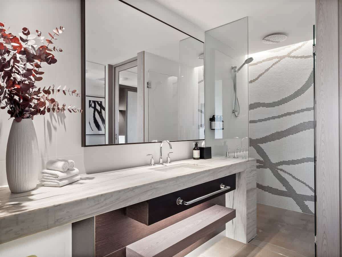 A modern bathroom with a marble countertop, a large mirror, a shower with a patterned glass door, and a vase with dried flowers.