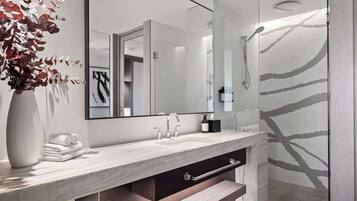 A modern bathroom with a marble countertop, a large mirror, a shower with a patterned glass door, and a vase with dried flowers.