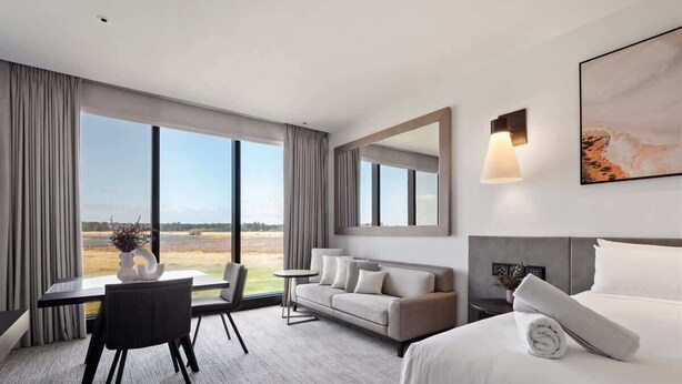 Une chambre d’hôtel comprenant un lit, un canapé, une table à manger avec des chaises, une petite table avec une plante et une grande fenêtre donnant sur un paysage.