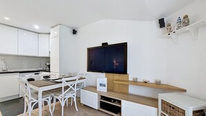 A modern kitchen with white cabinetry, a dining table with chairs, a flat-screen TV mounted on the wall, and a shelf with decorative items.