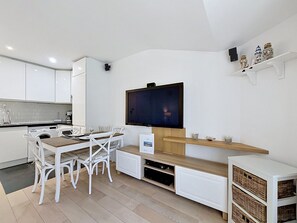 Una cucina moderna con mobili bianchi, un tavolo da pranzo con sedie, una TV a schermo piatto montata a parete e uno scaffale con oggetti decorativi.