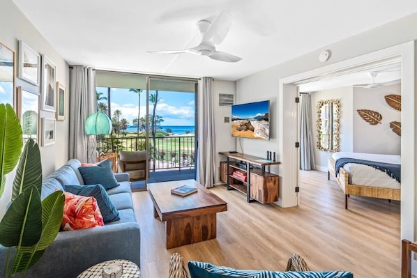 A modern living room with a sofa, coffee table, and TV, a balcony with a view, and a bedroom with a bed and curtains.