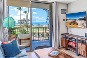 A living room with a sofa, a coffee table, a TV on the wall, and a balcony with a view of the ocean.