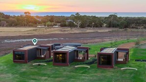 A modern outdoor setting with several square, glass-walled structures, likely guest rooms, arranged on a grassy area with a view of a natural landscape and a body of water in the background. - Samphire Chalet Tuart—Luxury Amidst Nature (Busselton)