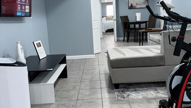 A modern living room with a treadmill, a flat-screen TV mounted on the wall, a dining area with a table and chairs, and a bedroom visible through an open door.