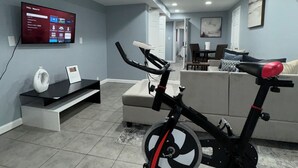 A spacious living area with a large flat-screen TV mounted on the wall, a stationary exercise bike, a sofa, and a dining table.