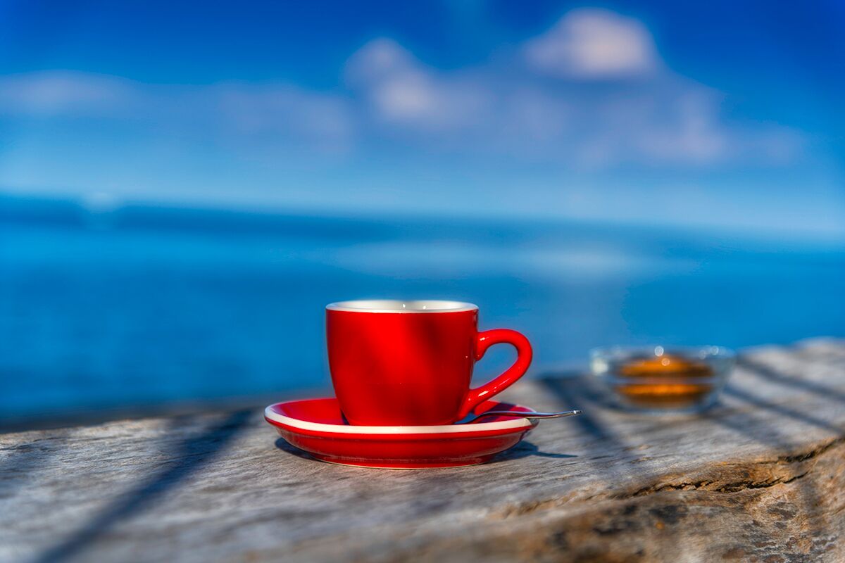 A red cup and saucer on a wooden surface with a blurred background of a blue body of water and a distant structure.