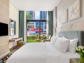 Hotelkamer met een groot bed, een tv aan de muur, een klein tafeltje met een stoel, een raam met uitzicht en een bloemenvaas op het nachtkastje.