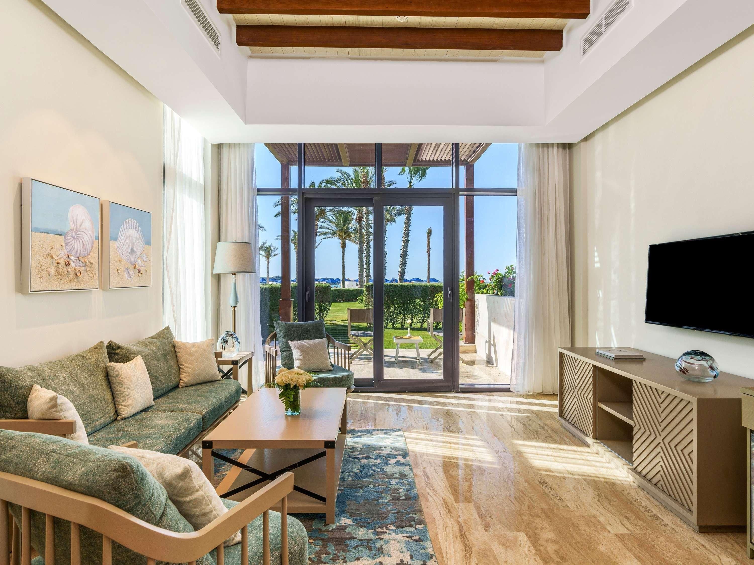 Una sala de estar moderna con sofá, mesa de centro y televisor, con un amplio ventanal que da a un balcón con vistas a palmeras y al mar.