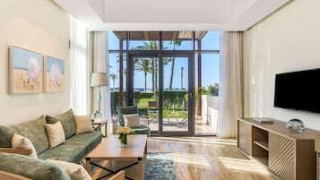 Una sala de estar moderna con sofá, mesa de centro y televisor, con un amplio ventanal que da a un balcón con vistas a palmeras y al mar.