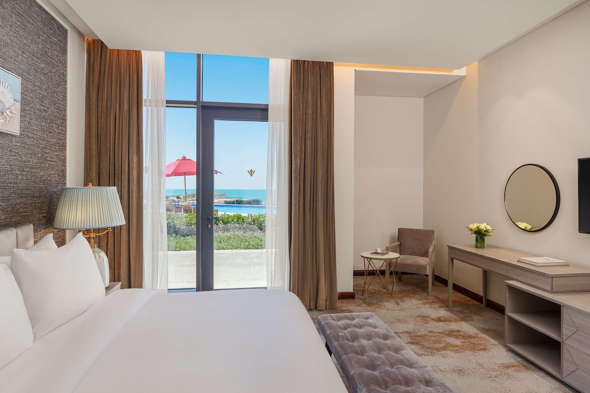 Une chambre d’hôtel avec un grand lit, un bureau, une chaise, une petite table avec une tasse, et une vue sur l’océan par une porte vitrée.