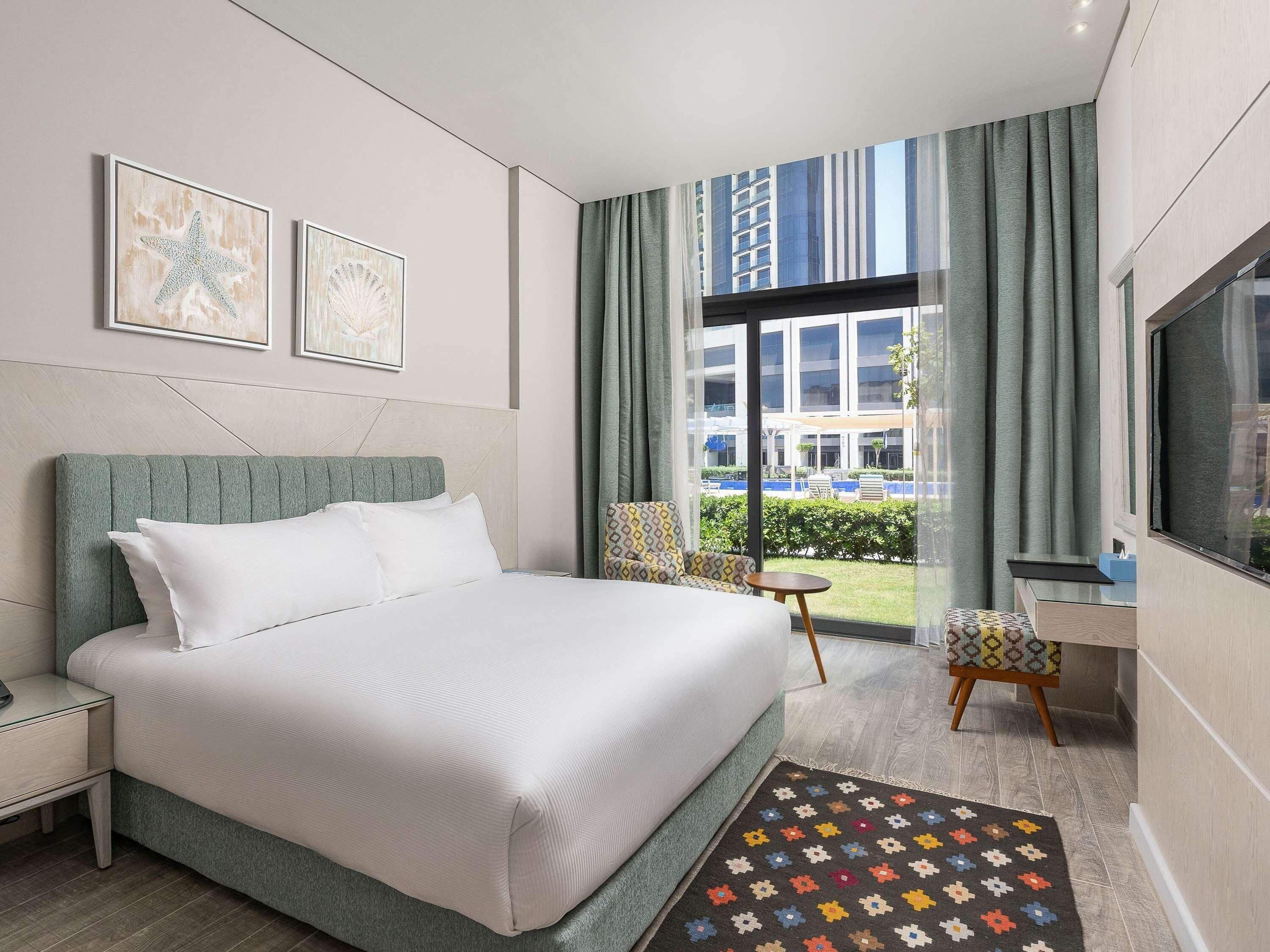 Ein modernes Hotelzimmer mit einem großen Bett, einem Schreibtisch, einem Sessel und Blick auf die Stadt durch große Fenster.