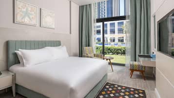 Ein modernes Hotelzimmer mit einem großen Bett, einem Schreibtisch, einem Sessel und Blick auf die Stadt durch große Fenster.