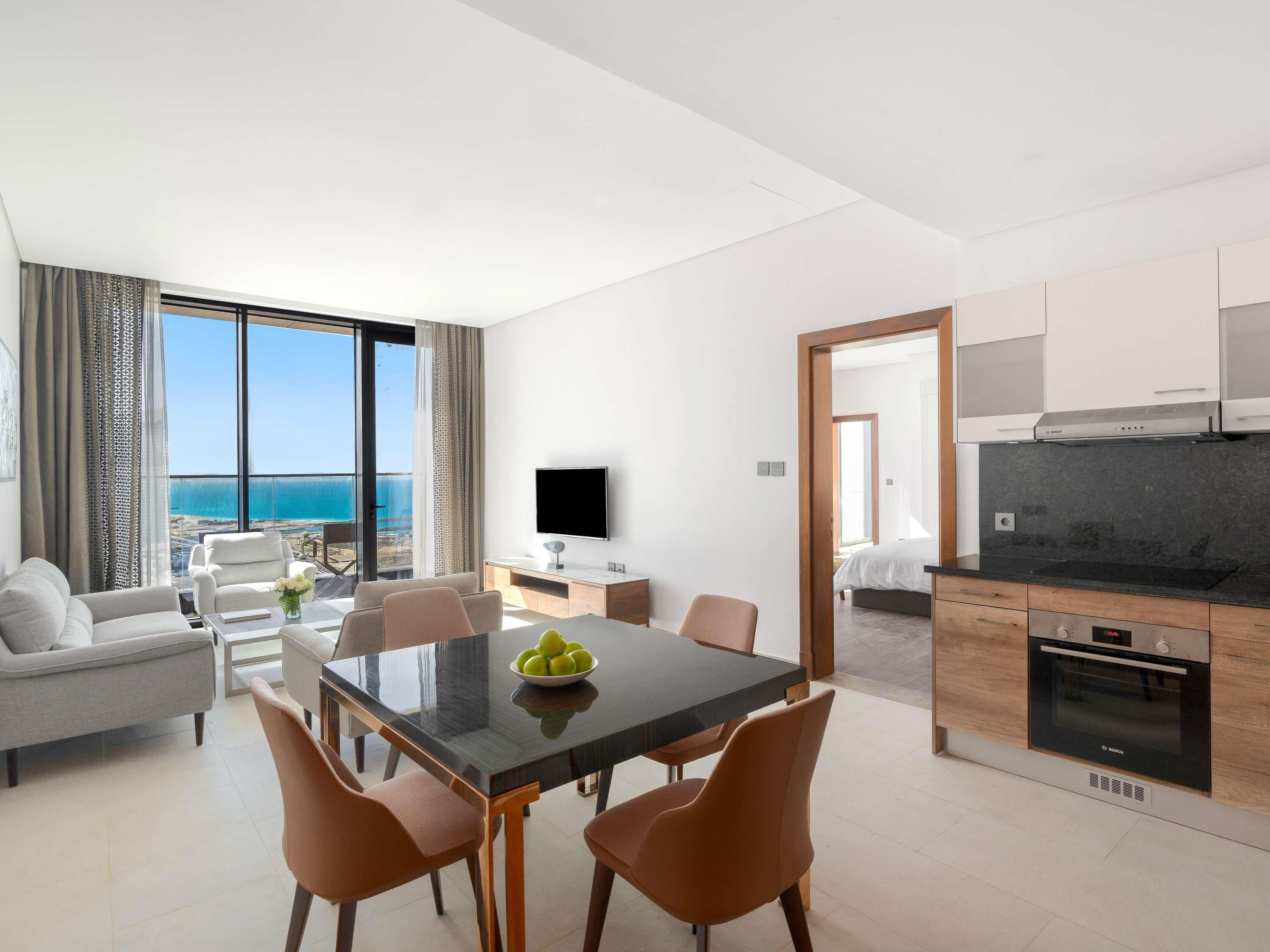 A modern hotel room with a dining area, a kitchen, and a large balcony with a sea view.