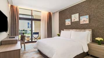 A modern hotel room with a large bed, a desk, and a chair. There are two windows with curtains, a view of the outside, and a wall with framed pictures.