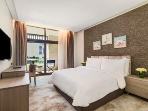 A modern hotel room with a large bed, a desk, and a chair. There are two windows with curtains, a view of the outside, and a wall with framed pictures. - Rixos Premium Alamein (El Alamein)