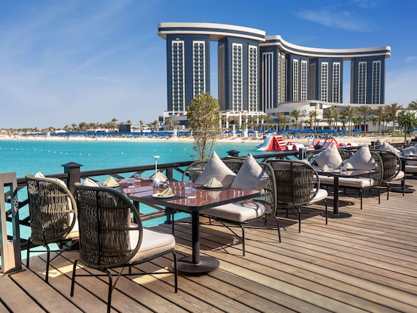 A seaside restaurant deck with outdoor seating, overlooking a beach and a hotel in the background. - Rixos Premium Alamein (El Alamein)