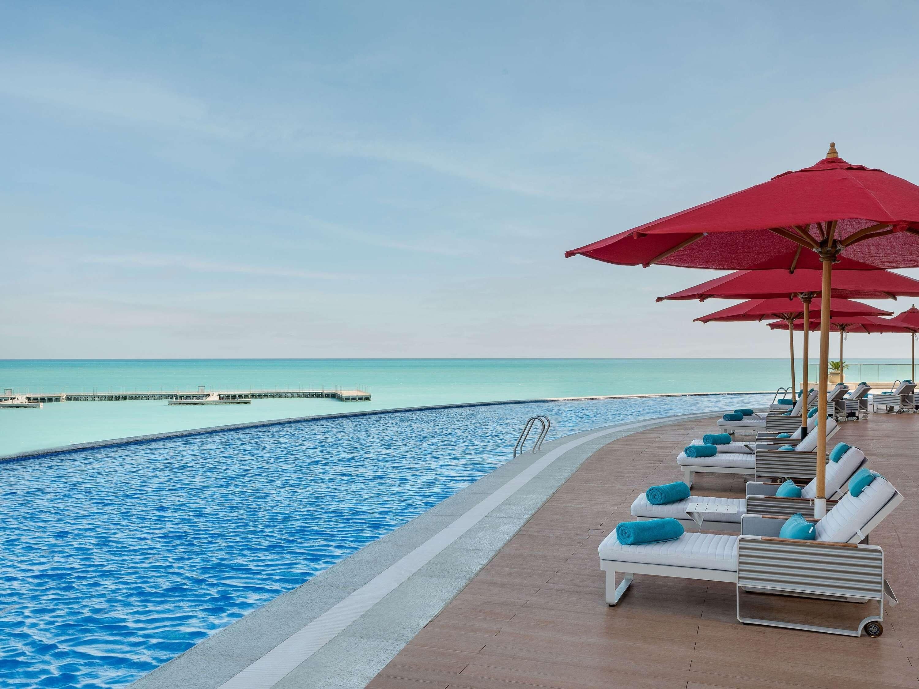 A poolside area with lounge chairs and red umbrellas, overlooking a clear blue sea and a pier.