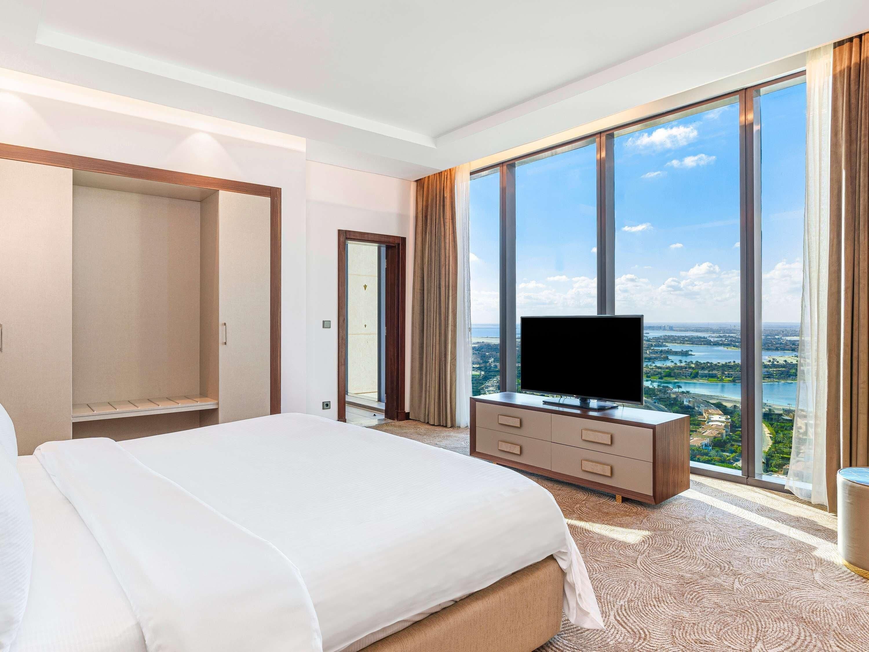 Une chambre d’hôtel avec un grand lit, un téléviseur posé sur une commode en bois et une vue sur le paysage urbain, visible à travers de grandes fenêtres.