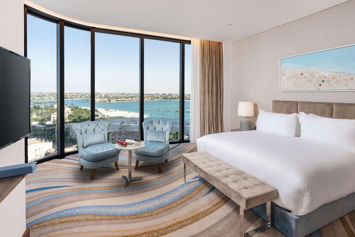 A hotel room with a large window, a bed, a bench, two armchairs, a small table, and a painting.