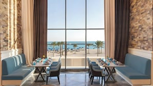 A dining area with a view of the beach, featuring a long table with chairs, cushioned seating, and a large window. - Rixos Premium Alamein (El Alamein)