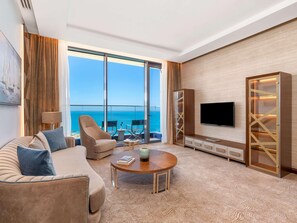 A modern hotel room with a large window offering a sea view, a comfortable sofa, a coffee table, and a TV on the wall.