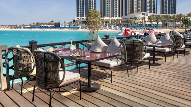 A seaside restaurant deck with outdoor seating, overlooking a beach and a hotel in the background. - Rixos Premium Alamein (El Alamein)