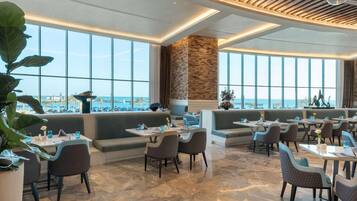 Un restaurant moderne offrant une vue sur la mer, avec un sol en pierre, des sièges confortables et un éclairage combinant des sources naturelles et artificielles.
