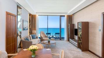 A hotel room with a large window offering a sea view, a dining area with a wooden table, and a seating arrangement including a sofa and armchairs.