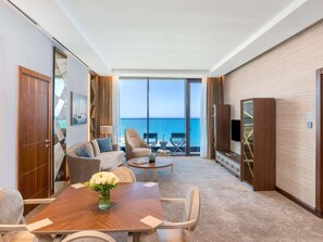 Een hotelkamer met een groot raam en uitzicht op zee, een zithoek met een houten tafel en een zithoek bestaande uit een bank en fauteuils.