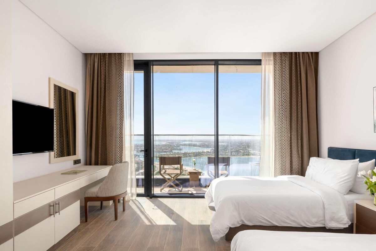Ein Hotelzimmer mit großem Fenster, einem Bett, einem Schreibtisch und einem Fernseher.