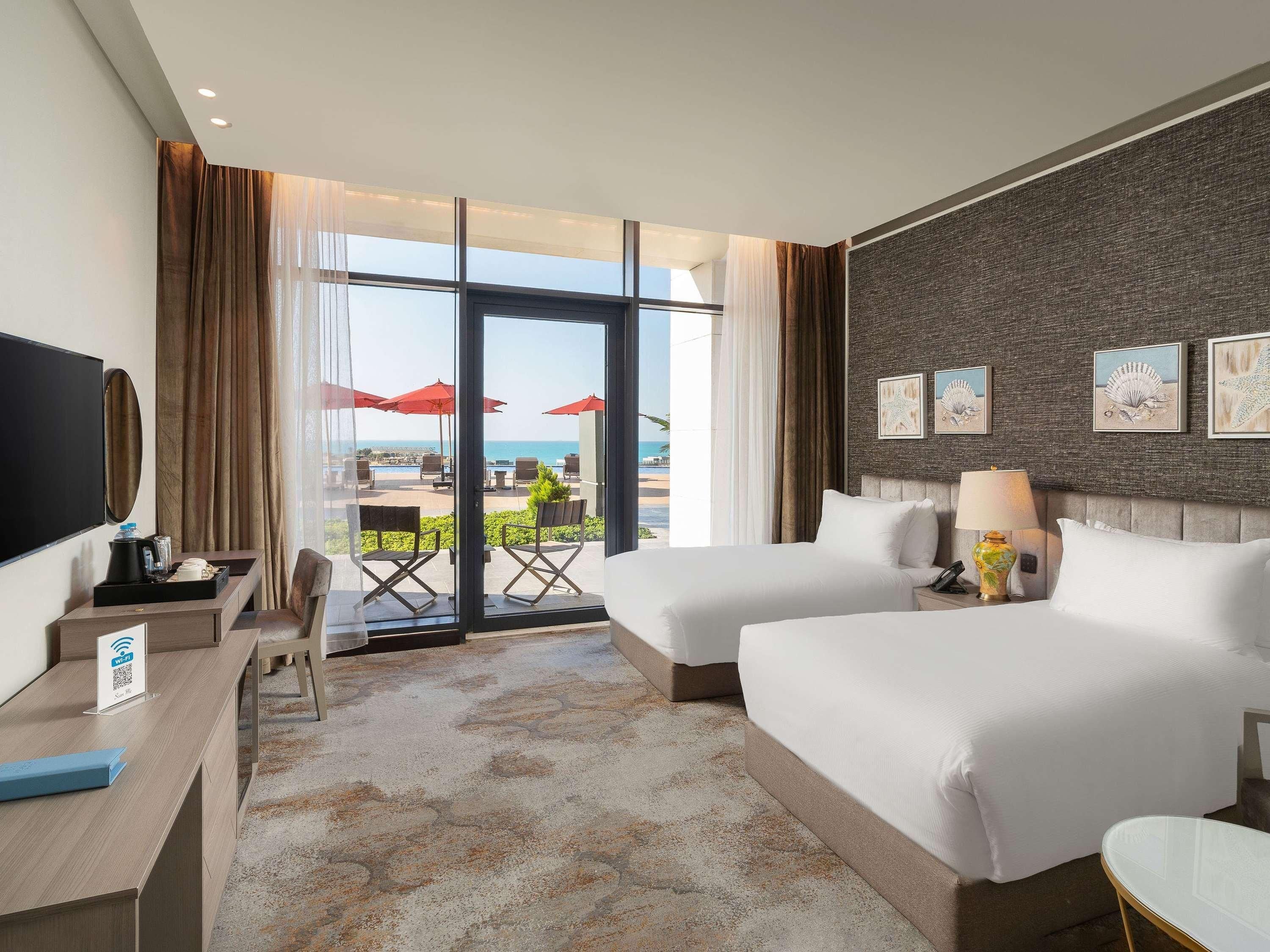 A hotel room with two beds, a desk, a TV, and a balcony with red umbrellas.