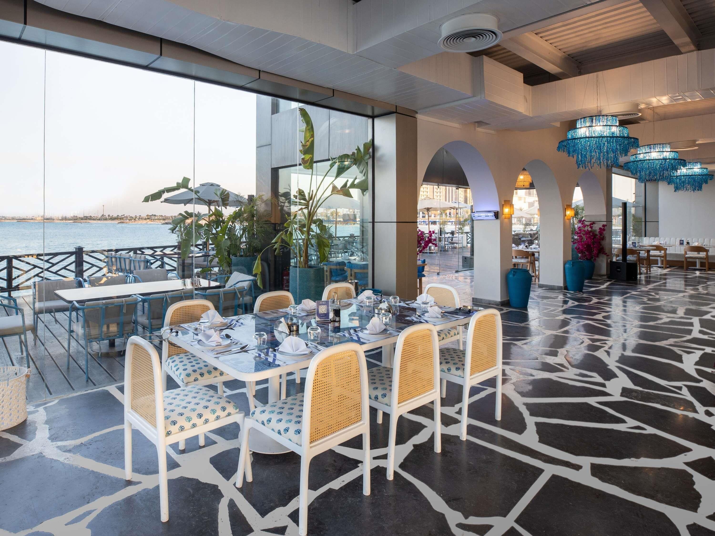 A restaurant with outdoor seating overlooking the sea, featuring white wicker chairs, glass tables, and blue hanging lights.