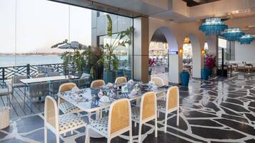 A restaurant with outdoor seating overlooking the sea, featuring white wicker chairs, glass tables, and blue hanging lights.